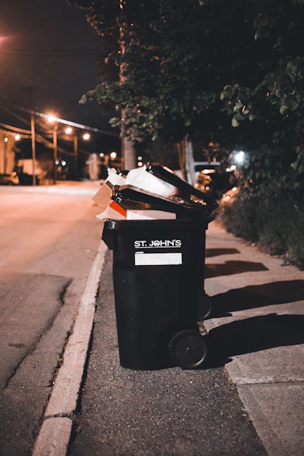 A black wheelie bin positioned on the pavement beside a residential street at night, with the label 'ST. JOHN'S' visible on the front. The lid of the bin is open, revealing various waste materials, including cardboard and plastic items, some of which are protruding from the top. The surrounding environment features dim street lighting, casting shadows on the asphalt and adjacent pavement, with trees and bushes lining the side of the road. In the background, faint streetlights and nearby parked cars are visible, creating a quiet, suburban ambiance. The scene suggests an instance of private waste disposal, potentially managed by local waste collection services such as Waste Collection Roehampton, aligning with on-site or alternative rubbish removal practices outside of standard street collection schedules.