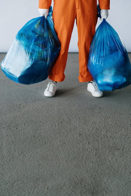 A person standing on a grey indoor floor, wearing orange work trousers and white sneakers, holding two large blue plastic refuse bags filled with waste in each hand. The bags appear to be made of durable, semi-transparent plastic, showing some contents inside. The background is plain with a white wall, suggesting an indoor environment such as a warehouse or storage area. The scene relates to waste collection or rubbish removal, highlighting an alternative waste handling method that might involve private rubbish collection services like those offered by Waste Collection Roehampton. The image emphasizes the physical act of rubbish disposal, suitable for illustrating the process of on-site clearance or manual rubbish removal, consistent with professional waste management practices.