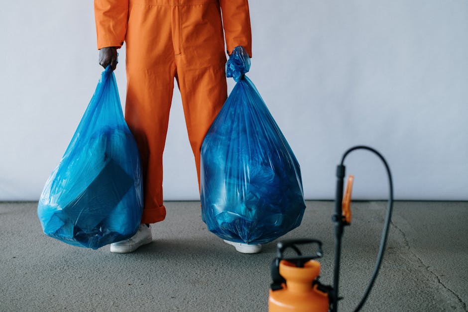 A person standing on a grey indoor floor, wearing orange work trousers and white sneakers, holding two large blue plastic refuse bags filled with waste in each hand. The bags appear to be made of durable, semi-transparent plastic, showing some contents inside. The background is plain with a white wall, suggesting an indoor environment such as a warehouse or storage area. The scene relates to waste collection or rubbish removal, highlighting an alternative waste handling method that might involve private rubbish collection services like those offered by Waste Collection Roehampton. The image emphasizes the physical act of rubbish disposal, suitable for illustrating the process of on-site clearance or manual rubbish removal, consistent with professional waste management practices.