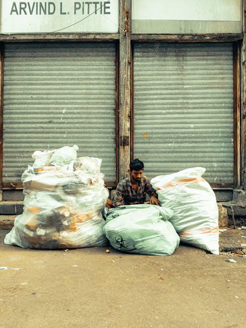 A man with dark hair and a patterned shirt is seated on the ground in front of a metal building with weathered, closed roller shutter doors. He is surrounded by several large plastic bags filled with mixed waste; some bags are semi-transparent, revealing content such as paper, plastic, and other debris, while others appear opaque and white with orange markings. The bags are positioned on a paved surface that appears slightly dusty, with small litter scattered nearby. The background shows a rusted metal frame supporting the shutters, and a partially visible sign with the name 'ARVIND L. PITTIE' at the top. The scene has a utilitarian, urban atmosphere, typical of informal waste collection or on-site rubbish clearance, consistent with alternative waste handling services offered by companies such as Waste Collection Roehampton. The lighting is natural, typical of overcast conditions, emphasizing the textures of the waste bags, the rough pavement, and the aged metal backdrop, all underlining the context of rubbish removal or disposal activities in a non-residential setting.