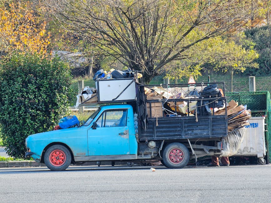 A small, older model flatbed truck painted in faded blue with red rims parked along the side of a street, loaded with various types of waste materials. The truck's cargo area contains a mixture of cardboard boxes, black plastic garbage bags, and loose debris, some of which are secured with ropes or tied to the sides. The materials appear to include paper, packaging, and refuse from a home or small business. A large, leafy green bush is visible near the front of the vehicle, partially obscuring the passenger side, while behind the truck there is a background of a tree with thin branches and green and yellow leaves, indicating an autumn setting. The environment suggests an on-site private waste collection or alternative rubbish removal service, characteristic of independent waste handling companies such as Waste Collection Roehampton, operating in residential or semi-urban areas. Overall, the scene exemplifies a typical rubbish removal scenario involving the loading of diverse waste materials for disposal or collection.
